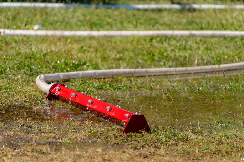 A red water curtain bar connected to a hose lies in a puddle on wet grass