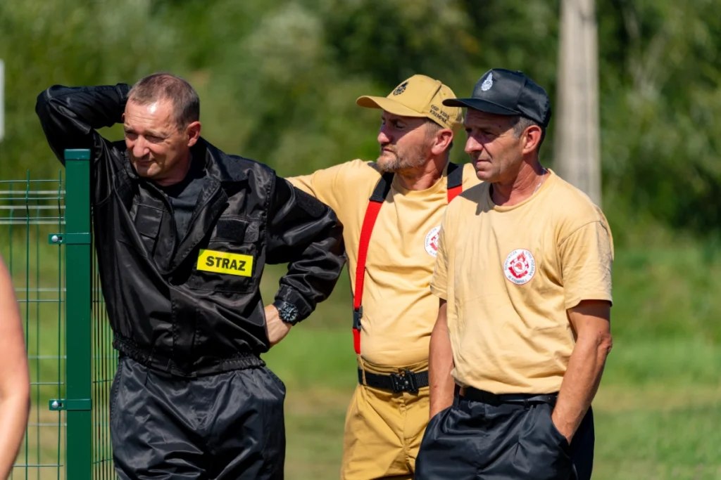 Three uniformed firefighters stand by a fence watching the demonstration, one wearing a STRAŻ patch