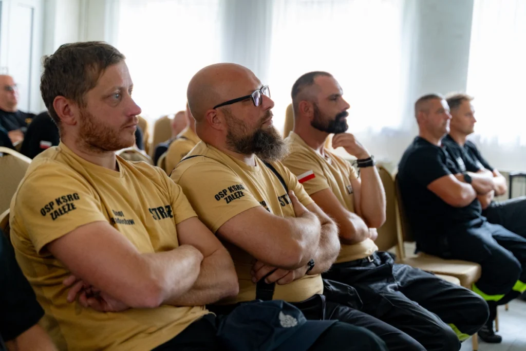 Volunteer firefighters in sand-coloured shirts reading OSP KSRG ZAŁEŻE listen to the training with arms folded