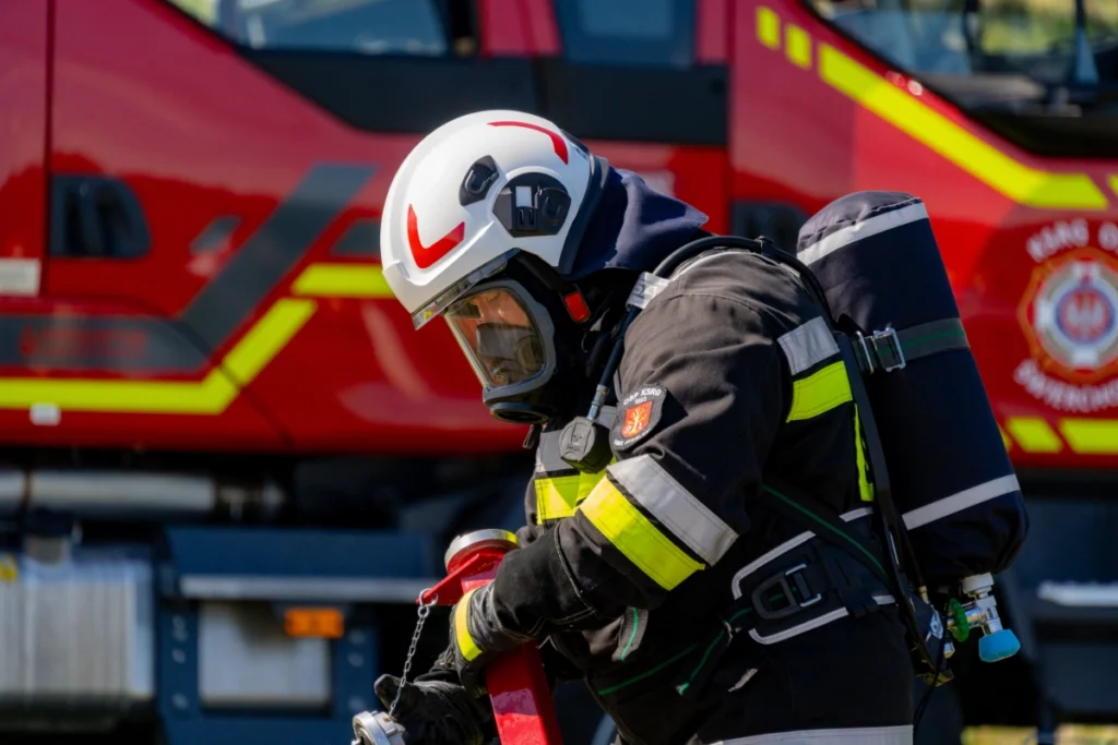 A firefighter in breathing apparatus, mask on, holds a red hose fitting in front of a fire engine