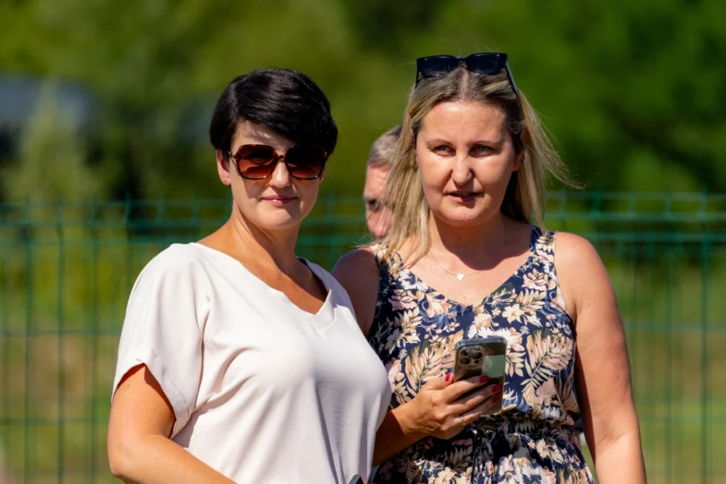 Two female picnic visitors stand by a green fence watching the demonstration, one holding a phone