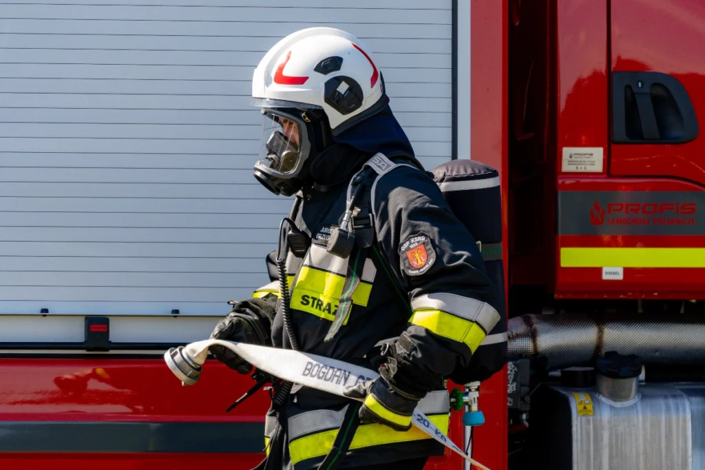 A volunteer firefighter in a white helmet and breathing apparatus holds a fire hose by a fire engine