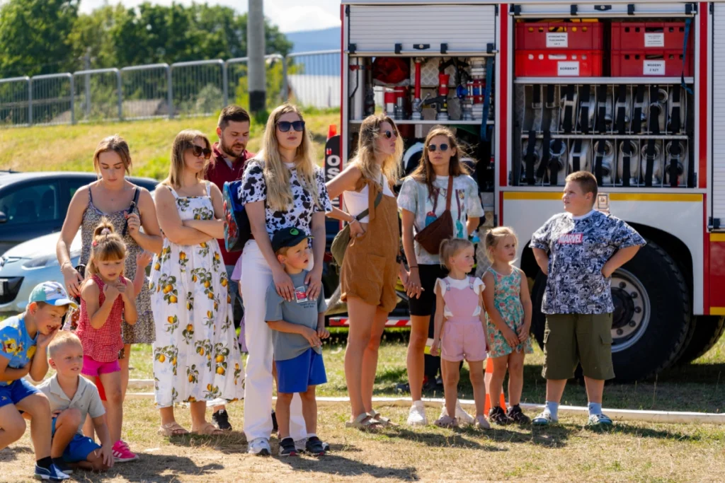 Families with children stand on the grass by a fire engine with open lockers, watching the demonstration