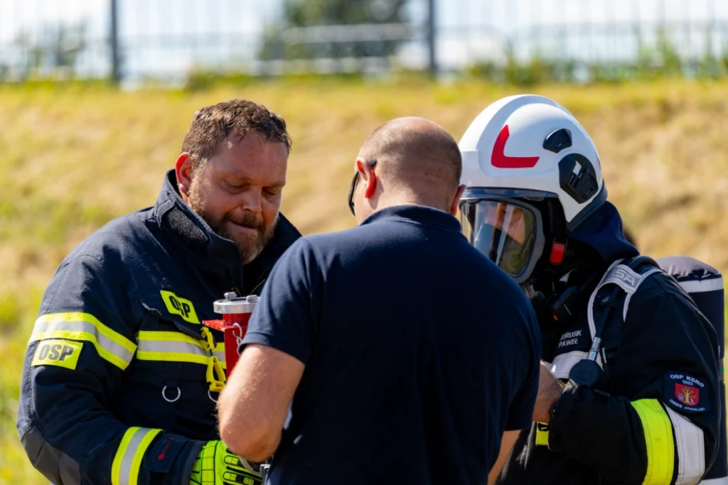 A man in a navy T-shirt talks with volunteer firefighters, one wearing a helmet and breathing apparatus