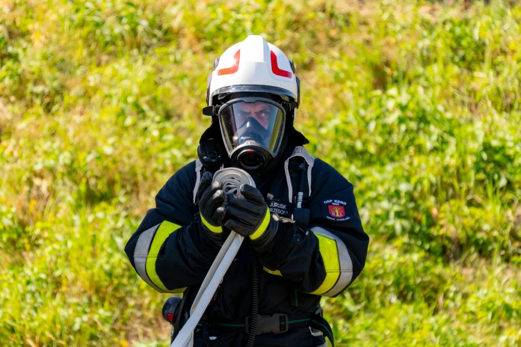 A volunteer firefighter in a white helmet and breathing apparatus mask holds a rolled-up fire hose