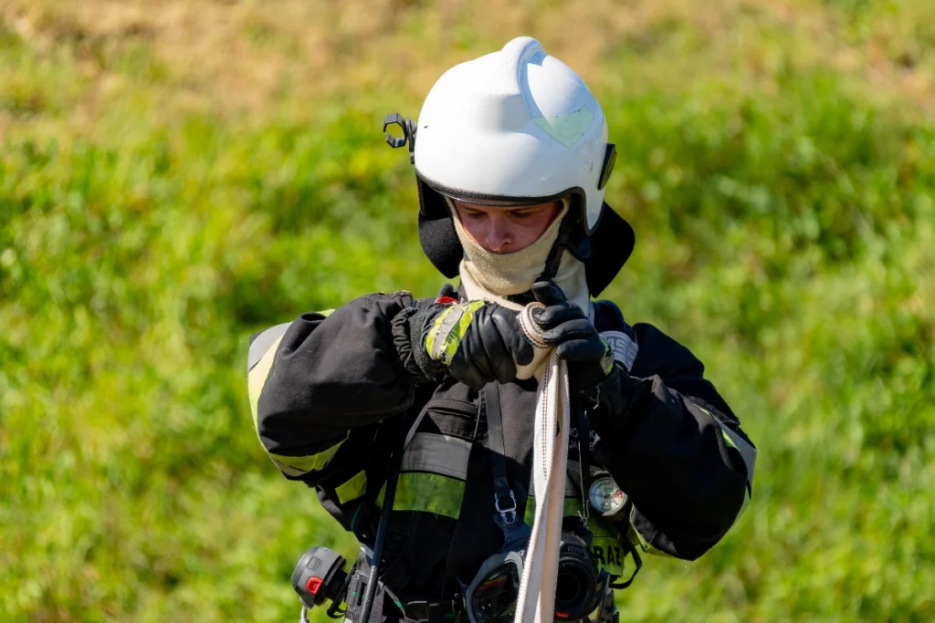 A white-helmeted volunteer firefighter in a balaclava rolls up a hose, breathing apparatus mask hanging on his chest