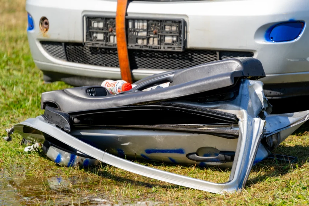 A silver car's cut-off door lies crumpled on the grass ahead of the car's front bumper
