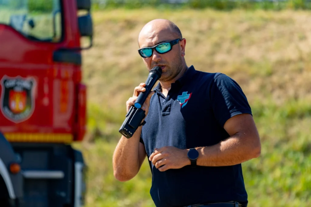 Training instructor in a navy shirt with the MED-POŻ logo speaks into a microphone beside a fire engine