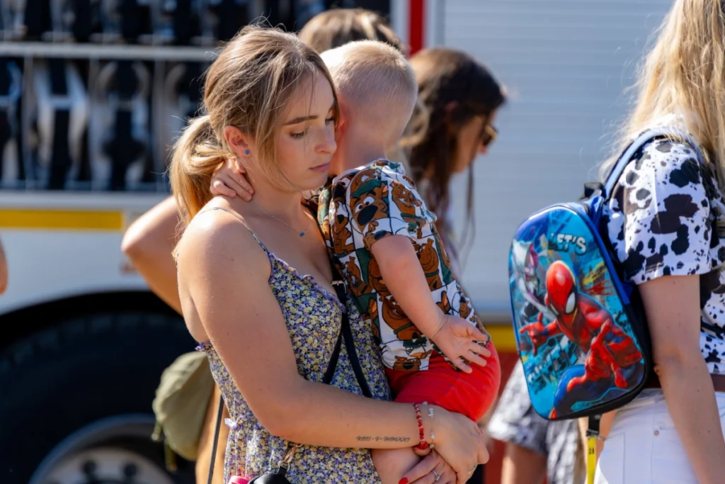 A woman holds a child with its arms round her neck, standing among spectators by a fire engine