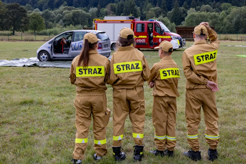 Czterech strażaków w mundurach z napisem STRAŻ, wśród nich dziecko, obserwuje pokaz ratownictwa