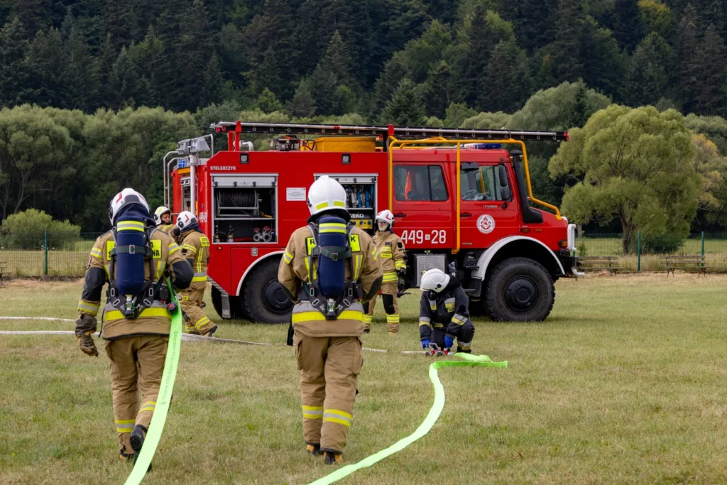 Strażacy idą z zielonym wężem gaśniczym w stronę wozu bojowego stojącego na boisku