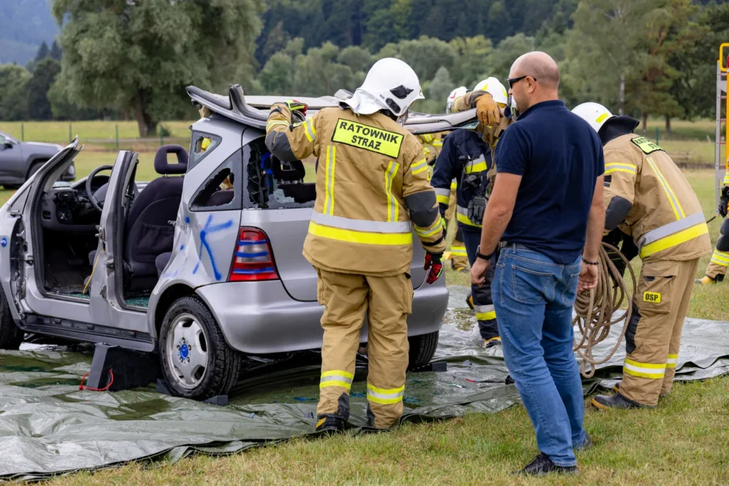 Strażak z napisem RATOWNIK STRAŻ zdejmuje odcięty dach samochodu, obok obserwują uczestnicy