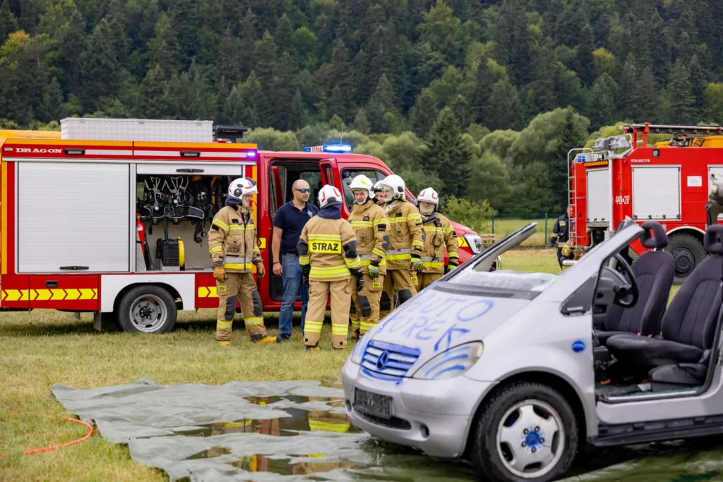 Strażacy rozmawiają przy otwartych skrytkach wozu bojowego, obok stoi rozcięty srebrny samochód
