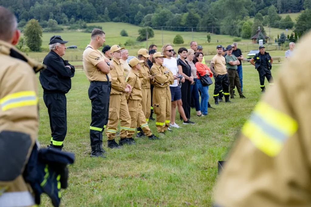 Strażacy, młodzież w mundurach OSP i mieszkańcy stoją na murawie, obserwując pokaz