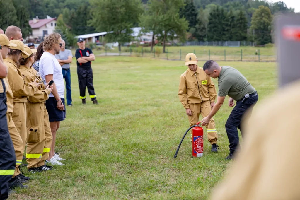 Instruktor przygotowuje gaśnicę razem z młodym strażakiem, uczestnicy przyglądają się z boku boiska