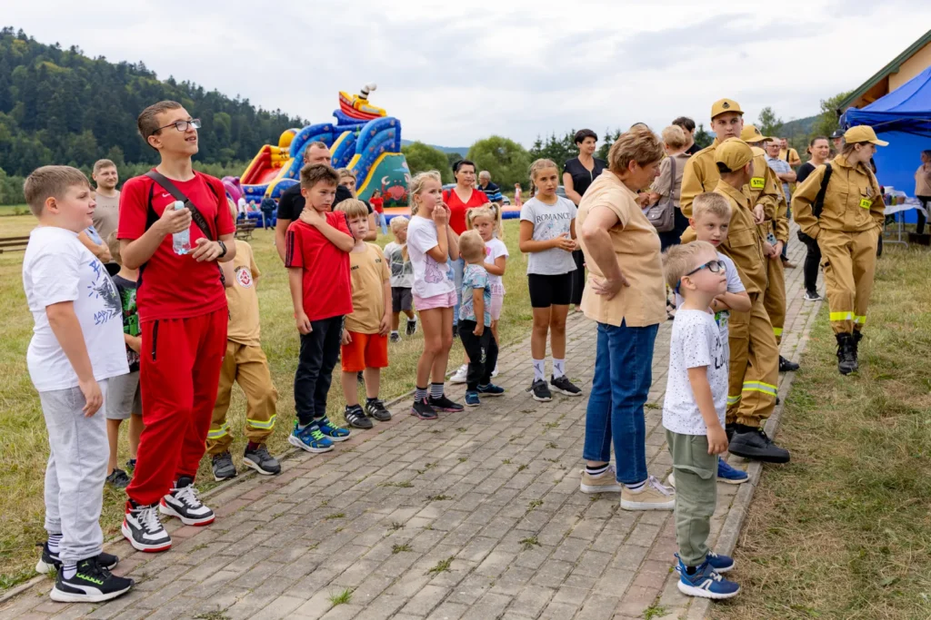 Dzieci i dorośli stoją wzdłuż chodnika i obserwują pokaz, obok młodzi strażacy