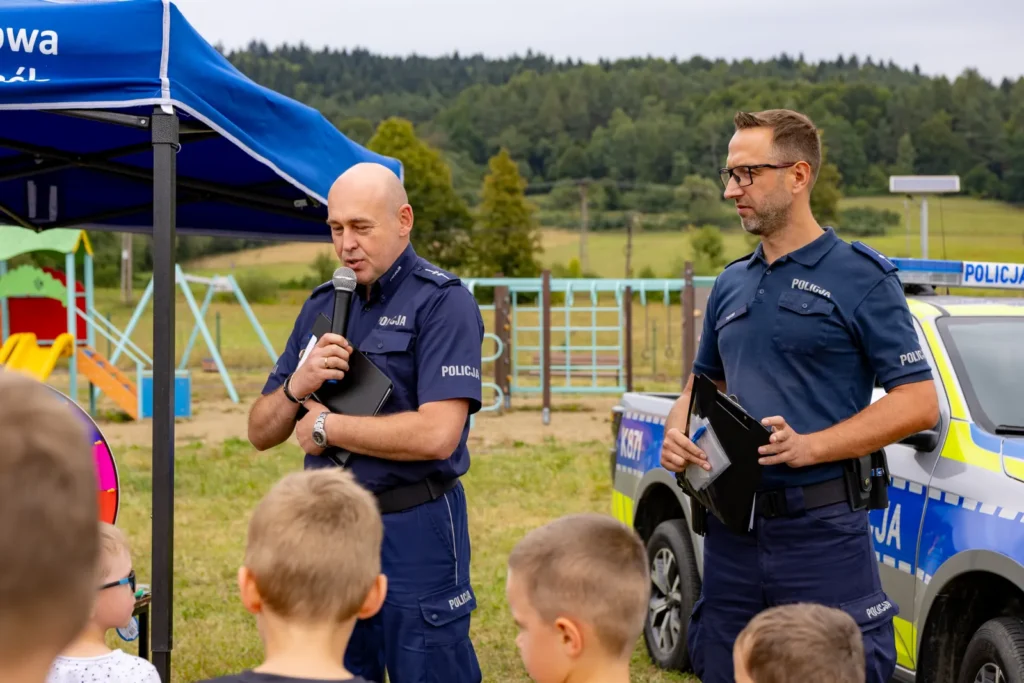 Policjant z mikrofonem mówi do dzieci przy namiocie, obok drugi funkcjonariusz i radiowóz
