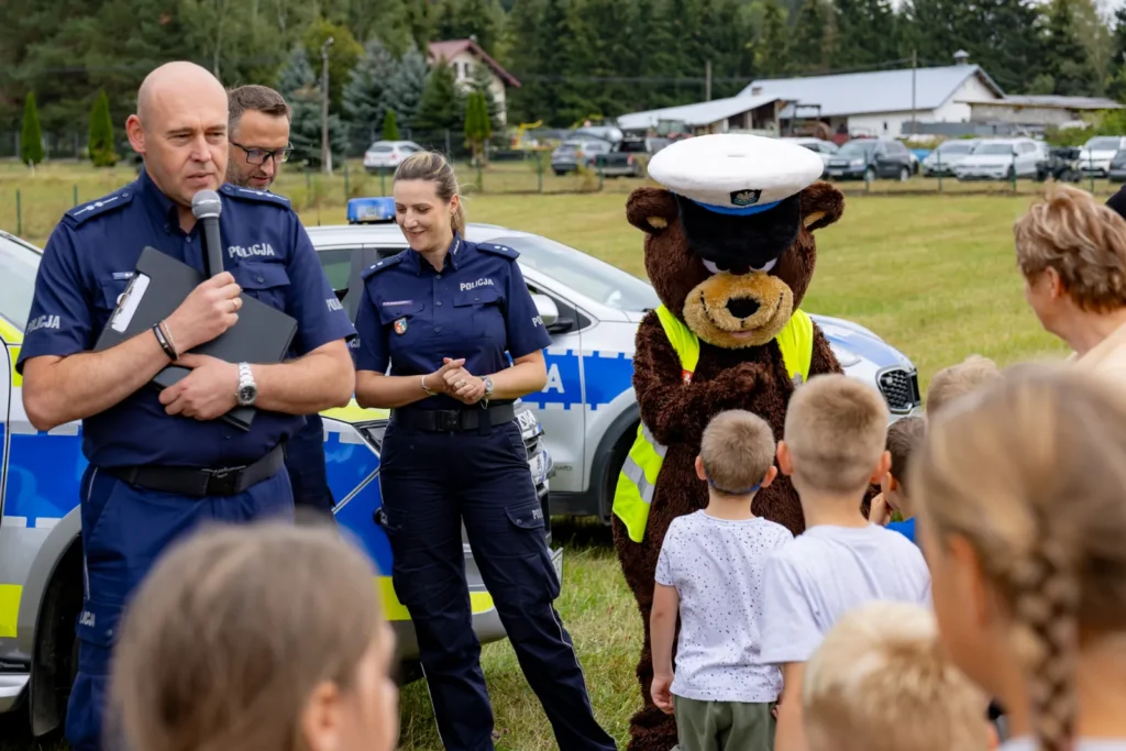 Maskotka policyjnego misia w kamizelce odblaskowej wita dzieci, obok trójka policjantów