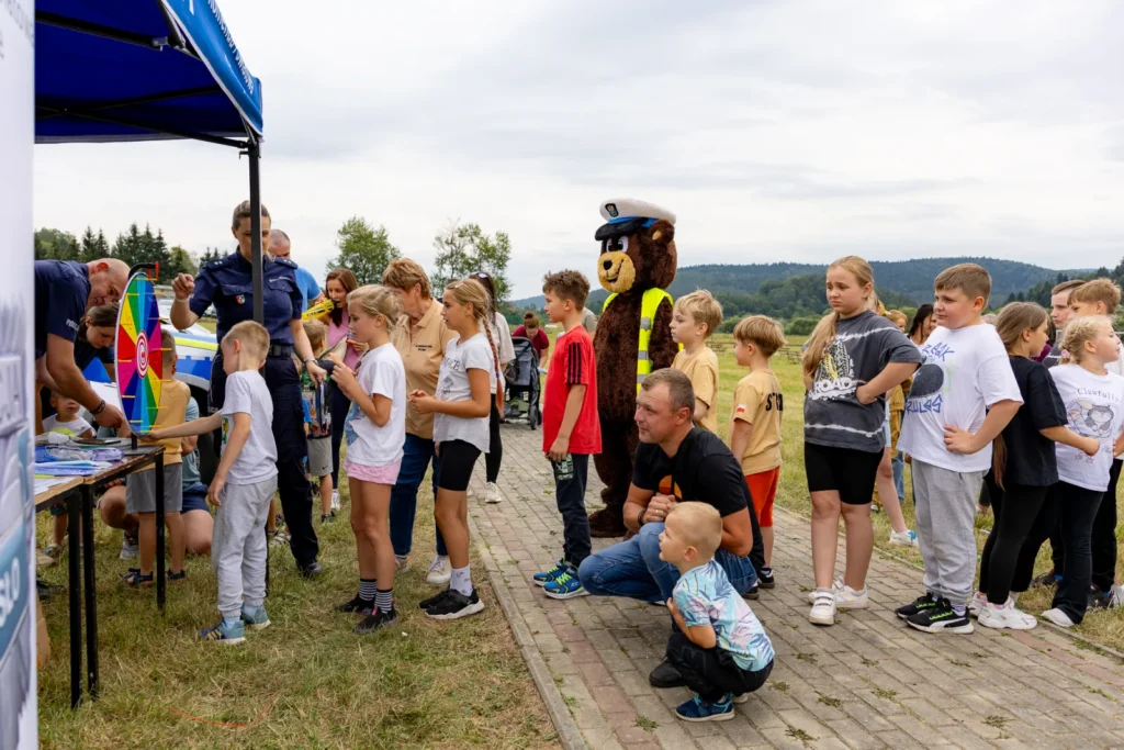 Dzieci w kolejce do policyjnego namiotu z kołem fortuny, obok maskotka misia