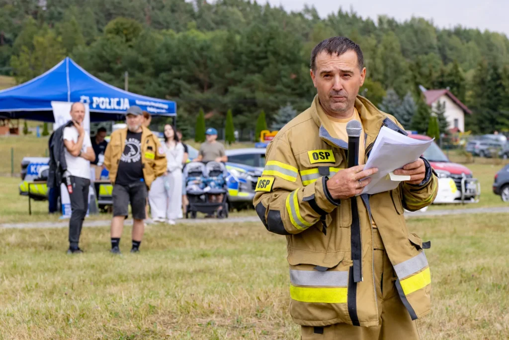 Strażak OSP z mikrofonem i kartkami mówi do uczestników, w tle namiot policji