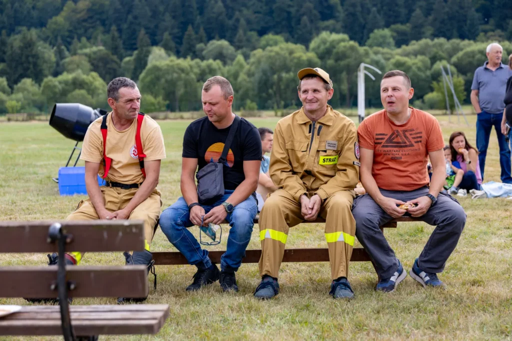 Czterej mężczyźni, w tym strażacy w mundurach, siedzą na ławce i oglądają pokaz